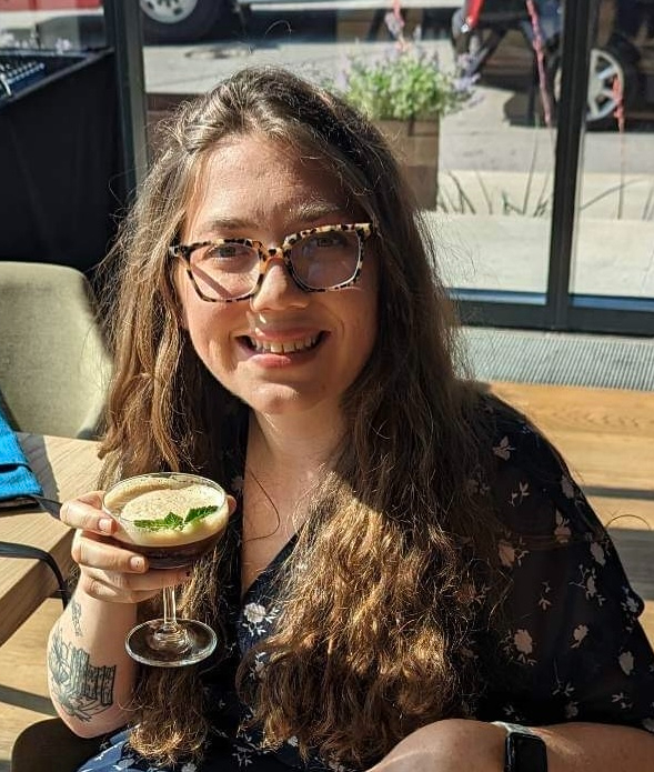 Gracie Bain headshot, young woman in glasses with long hair, holding a drink